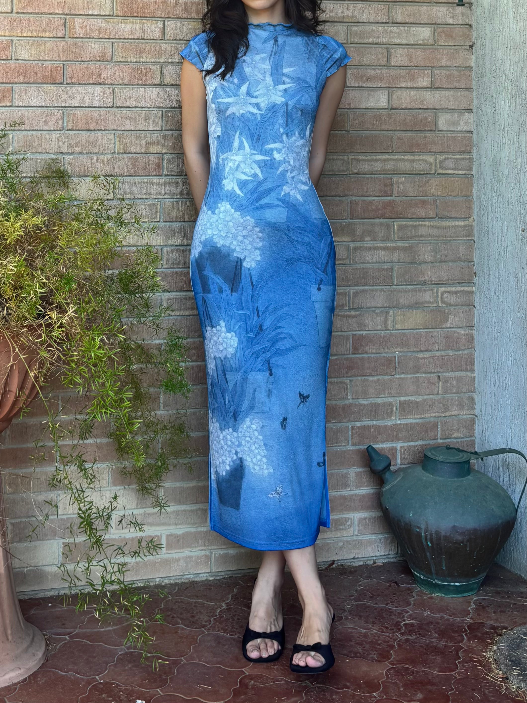 A woman wearing a blue cheongsam dress with a white floral print, standing against a brick wall.